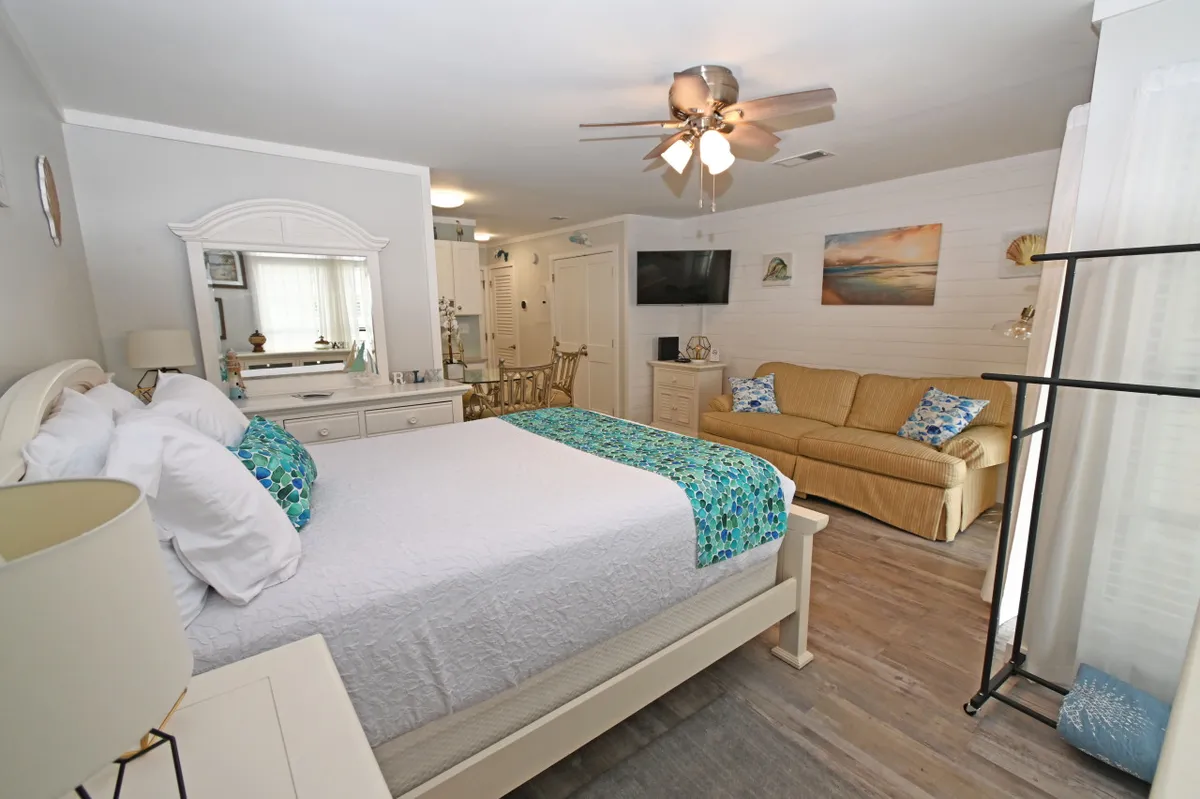 This image captures a sunlit studio interior featuring a plush queen-size bed adorned with colorful bedding, a comfortable sofa with decorative cushions, and a dressing table with a mirror. The arrangement feels open and inviting, with natural light enhancing the serene, cozy ambiance. Tasteful artwork on the walls and thoughtful décor elements complete the scene, emphasizing modern coastal comfort.