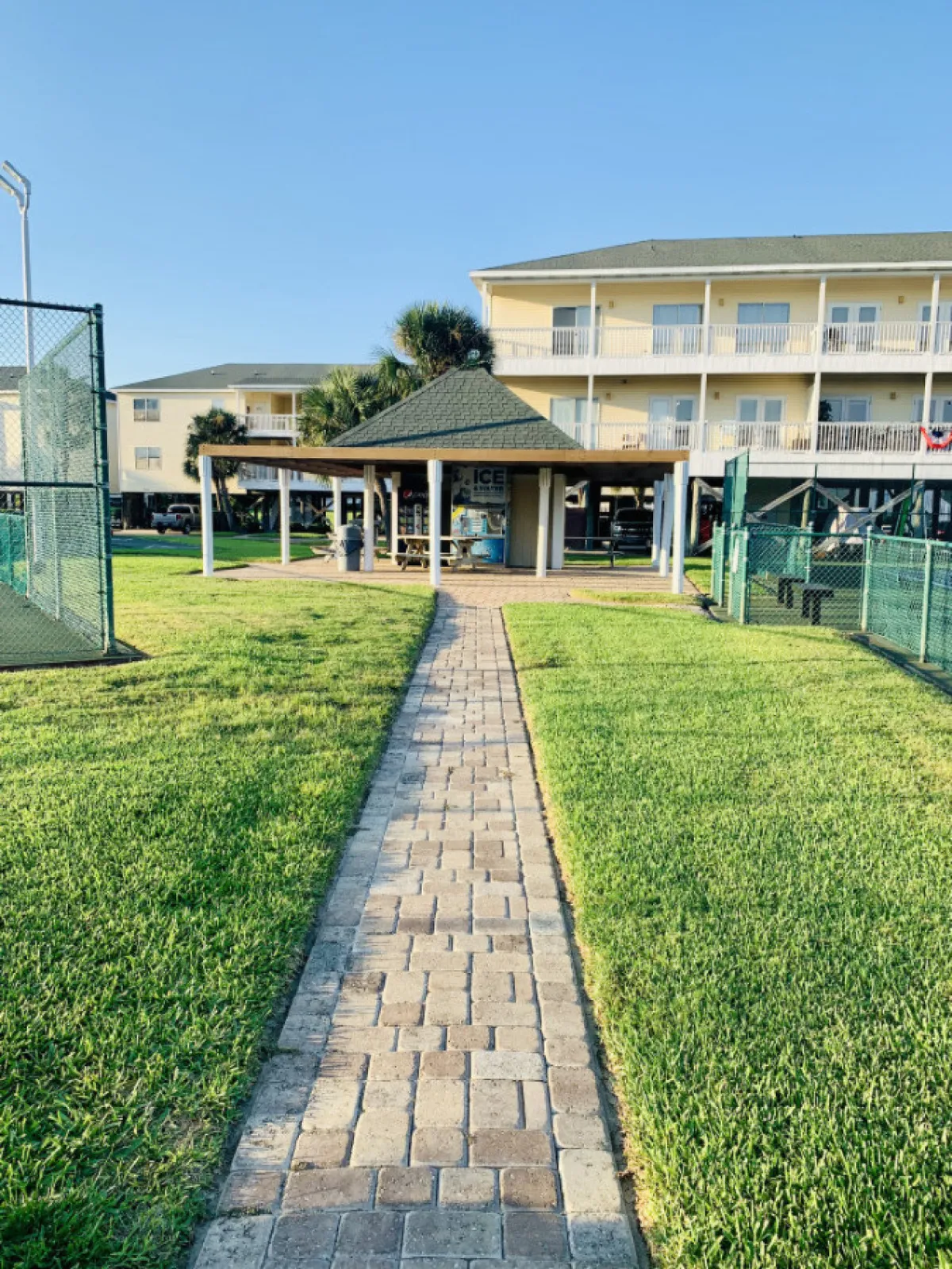 A cheerful pathway bordered by green grass leading to a gazebo beside a building. The scene is bright and welcoming, with a clear sense of community and inviting outdoor areas for socializing or relaxing.