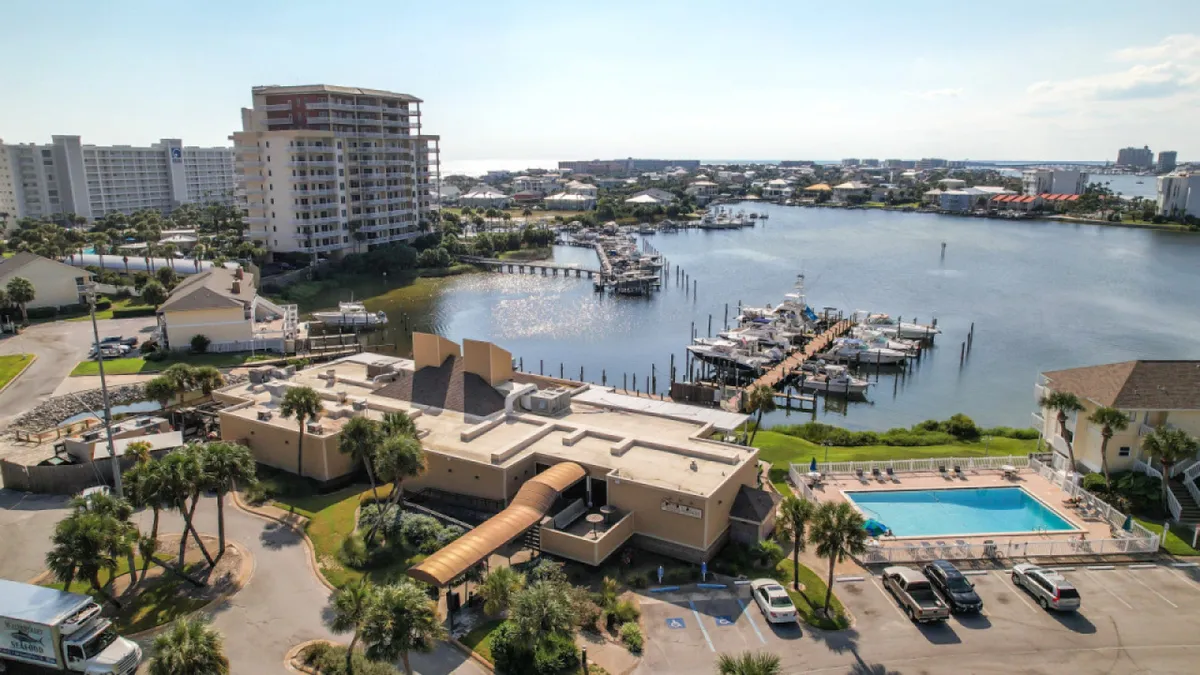A wide exterior shot of the property’s marina, swimming pool, and landscaped greenery, with residential buildings in the background. The scene is leisurely, offering glimpses of walkable paths and community gathering spots along the waterfront.