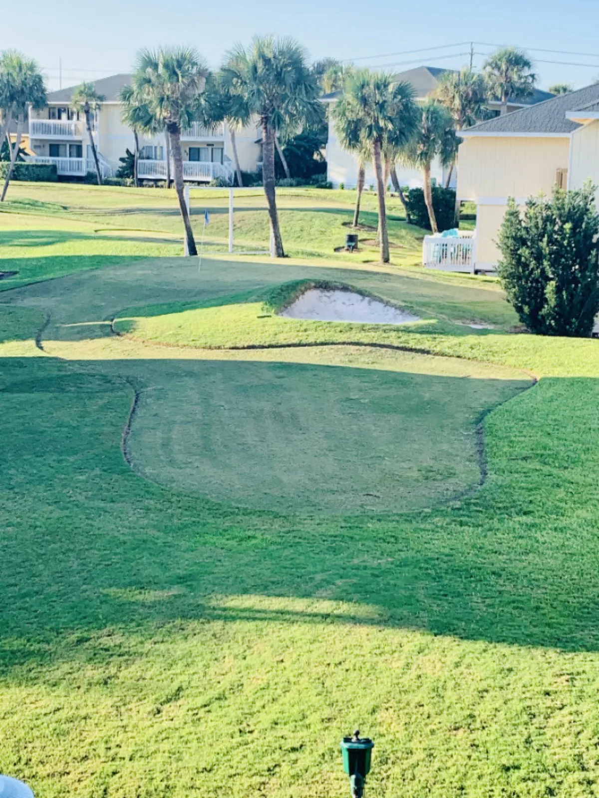 A sun-drenched view of the resort's 9-hole golf green, set among swaying palm trees and bordered by residential buildings. The sand trap and manicured greens are visible, inviting guests to tee off or enjoy a leisurely stroll outdoors. Groups of people can be seen golfing and socializing, with the overall atmosphere relaxed and friendly.