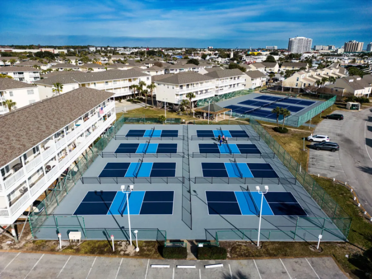 Energetic tennis courts are at the forefront, with people playing matches while others watch from the sidelines. The courts are framed by well-maintained green spaces and the resort’s residential buildings. The mood is lively, suggesting the vibrant community atmosphere where guests can participate in friendly competitions or casual games.