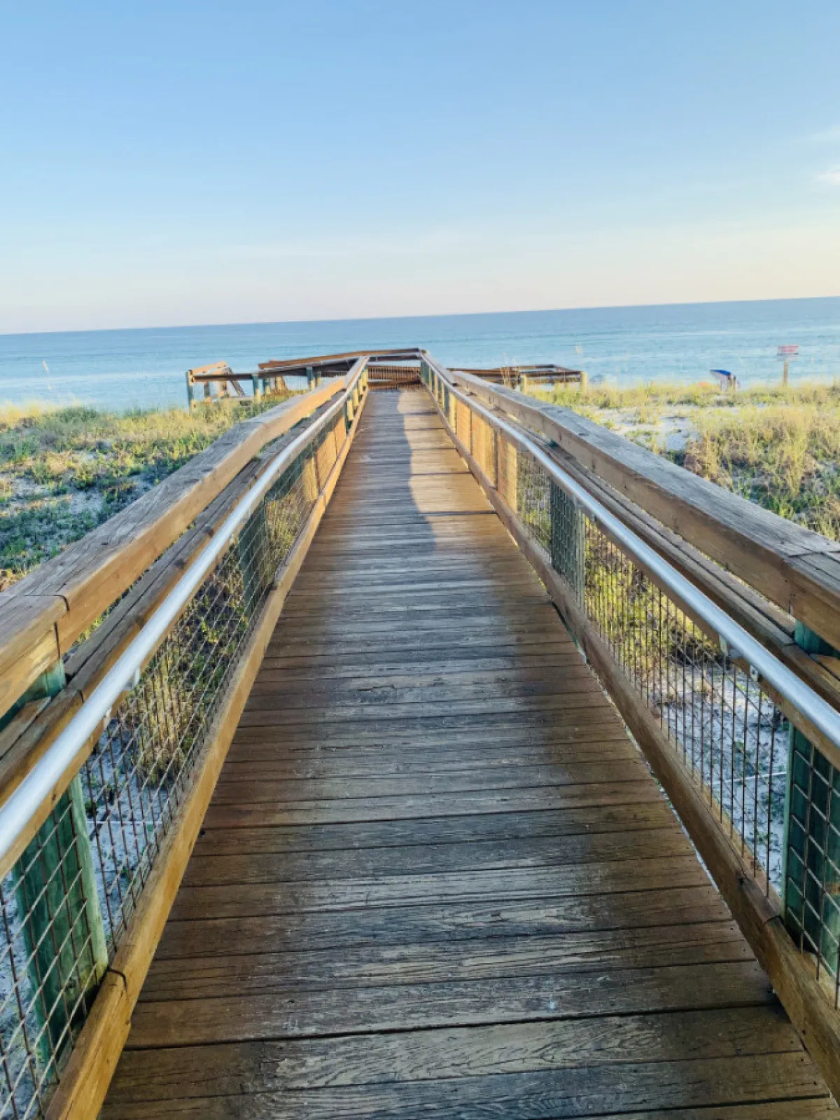 This image captures a wooden boardwalk leading directly toward the ocean, with expansive views of the blue water and distant piers. The sandy beach is visible, invoking the feeling of stepping out for a beach day. The sky is clear, and the atmosphere is calm and inviting, perfectly evoking the classic Destin beachfront experience.