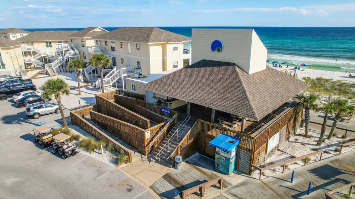 A beachfront property is shown with wooden walkways weaving through palm trees and a seating area that overlooks the sand. The scene highlights both the immediate access to the beach and welcoming communal spaces for relaxation or social gatherings, all set against the backdrop of soft dunes and the Gulf.