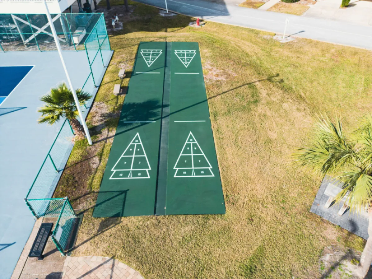 A wide view of shuffleboard courts surrounded by vibrant grass, palm trees, benches, and adjacent tennis courts. The open green space is perfect for walking, relaxing, and casual outdoor activities.