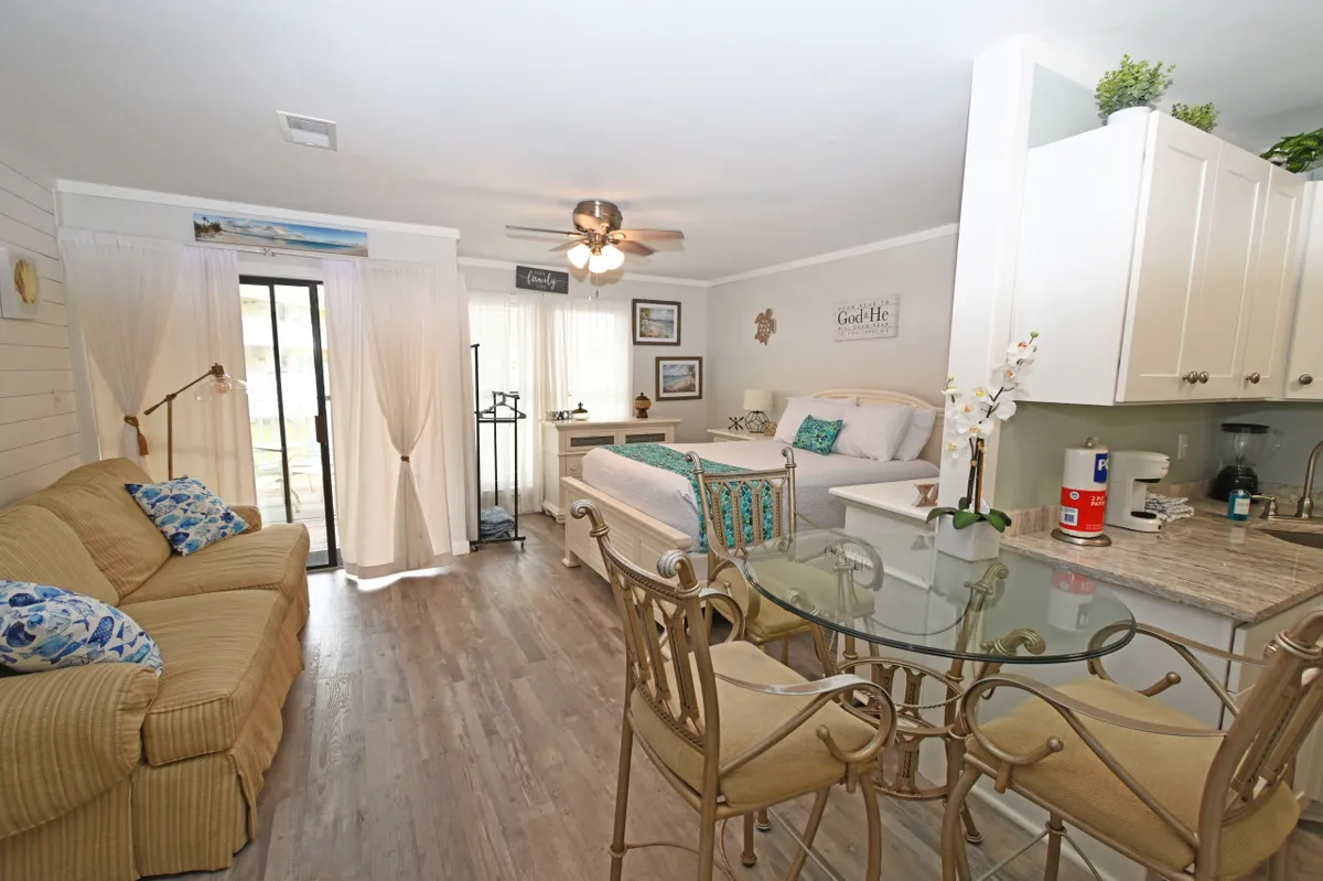 An inviting interior scene highlights a sofa with blue cushions, a bed with decorative pillows, and a glass dining table set within an airy, sun-filled space. The layout is open and functional, ideal for relaxing, dining, or winding down in style. Sunlight streams through light curtains, adding warmth and comfort to the modern coastal décor.