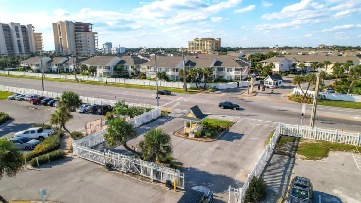 The entrance gate framed by palm trees and the main road, giving a welcoming impression of arrival and access to local attractions. The surrounding community buildings and lush greenery hint at the broader Destin neighborhood and its inviting atmosphere.