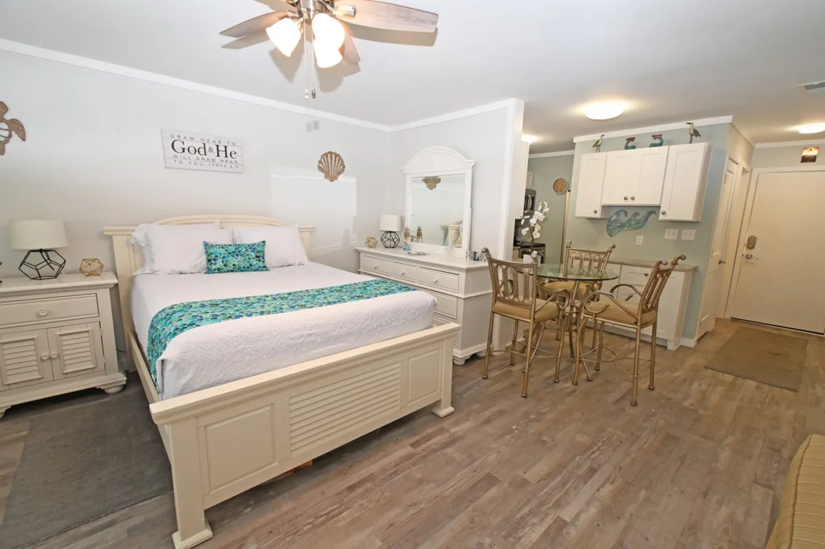 The image showcases a bright, airy studio with a queen-size bed dressed in turquoise and white bedding, a small dining table for four, a white dresser with a mirror, and a compact kitchenette. The decor is fresh and beach-inspired, creating a calm and inviting atmosphere perfect for relaxing after a day at the coast.