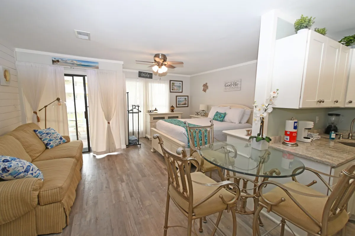 A bright, open-plan studio interior with a kitchenette featuring modern appliances and a granite countertop, a glass dining table with ornate chairs, a queen-size bed with decorative pillows, and a cozy sofa. The space exudes comfort and showcases the versatile, user-friendly kitchen and dining setup.