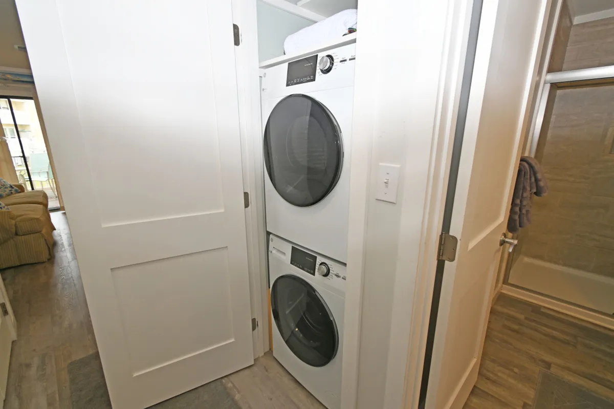 A compact yet functional laundry nook featuring stacked washer and dryer units just outside the bathroom. Doorways reveal the bathroom with its glass shower enclosure and a glimpse into the living area, demonstrating the seamless transition between utility and relaxation spaces.