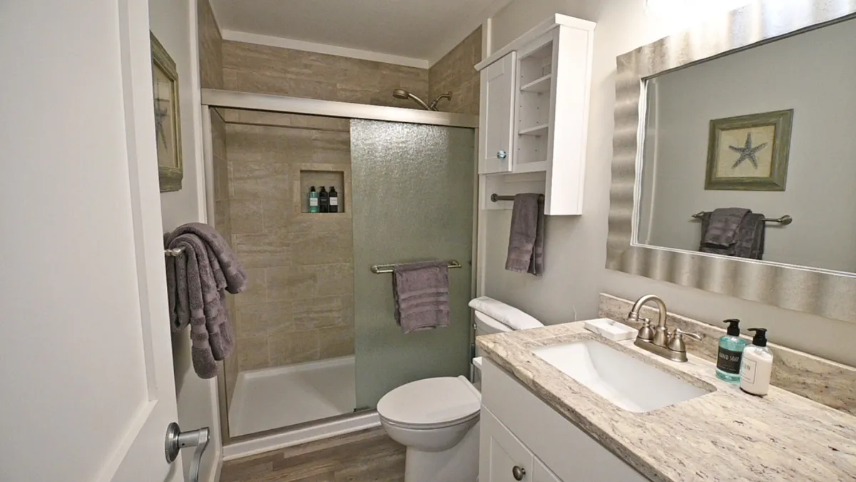 A modern, freshly renovated bathroom featuring a glass-enclosed shower with sleek tiled walls. The space is accessorized with a white vanity topped with a granite countertop, a large decorative mirror, and wall-mounted storage. Soft luxury towels and toiletries are neatly arranged, evoking a spa-like ambiance.