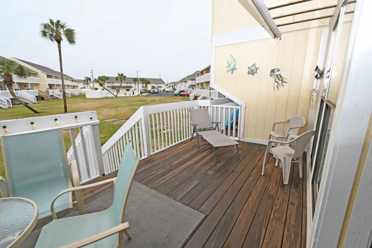 A wooden deck with cozy seating and marine-themed decor overlooks a green common area and neighboring buildings. White railings and stairs provide direct access to the outdoors, with clear sightlines to where pets might play. The tranquil mood is ideal for enjoying coffee or socializing, while watching pets in the grass below.