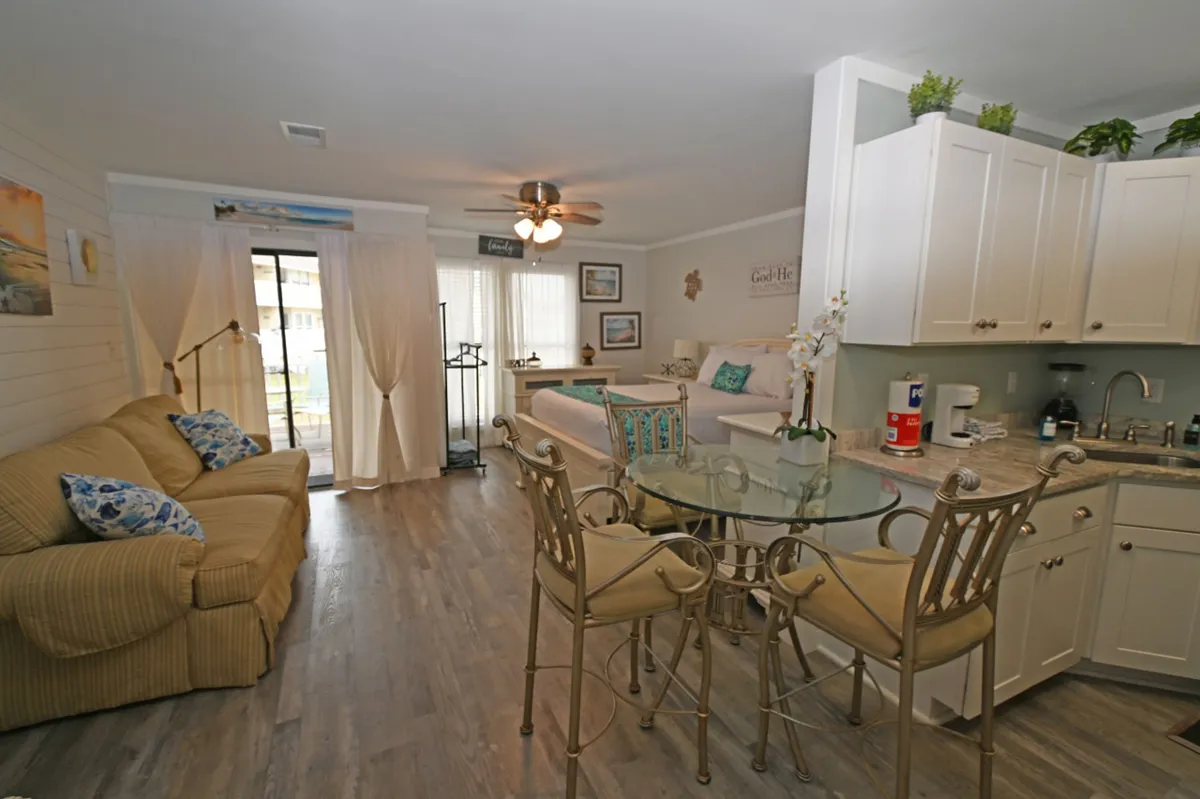 A stylish and welcoming studio interior featuring a comfortable queen-size bed with coastal pillows, a plush sofa with patterned cushions, a glass-top dining table with decorative chairs, and a compact, well-equipped kitchen with white cabinetry. Sliding glass doors hint at easy access to outdoor amenities and natural light fills the space, enhancing the sense of openness.