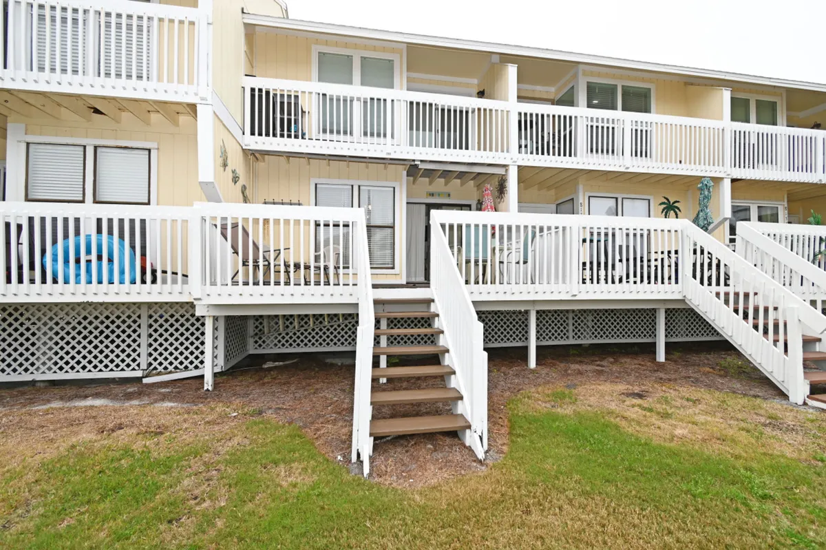 This image highlights the white wooden railings and stairs leading from an extended private deck down to a spacious, green grassy area in front of ground-level units. The yellow exterior walls with white trim evoke a cheerful coastal vibe. The scene features relaxing or dining on the patio, pets playing in the grass, and a sense of easy access to outdoor community spaces.