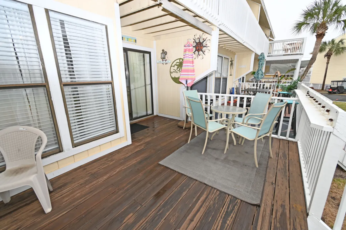 A cozy patio area with a table and four chairs, colorful beach towel and wall decorations, set on a wooden deck with white railing. The outdoor space connects seamlessly to the rest of the community and is ideal for relaxing, dining, or letting pets enjoy fresh air and sunshine right outside the door.