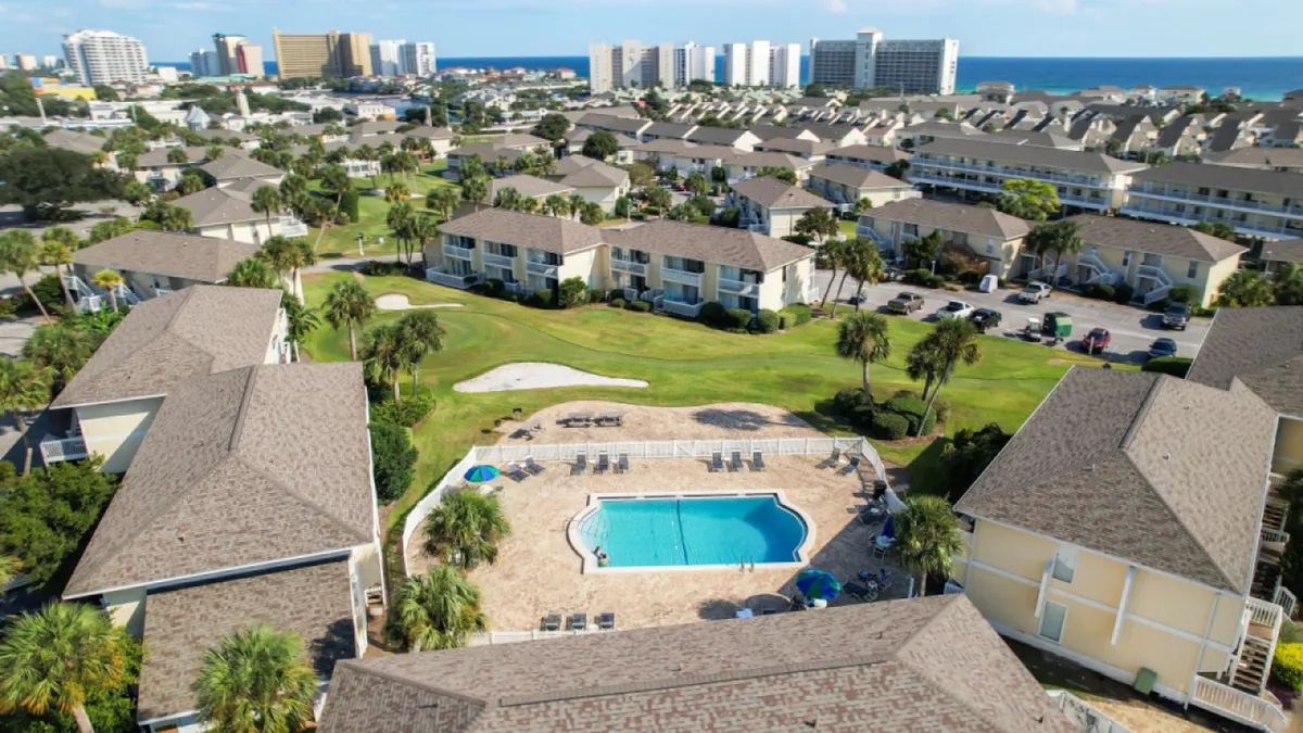 A central swimming pool surrounded by lounging chairs, lush green spaces, and palm trees, with clusters of low-rise condominium buildings in the background. The distant Gulf of Mexico and beachfront high-rises are visible, suggesting close proximity to the beach and a vibrant coastal environment. The scene is lively and welcoming, ideal for families enjoying resort amenities.