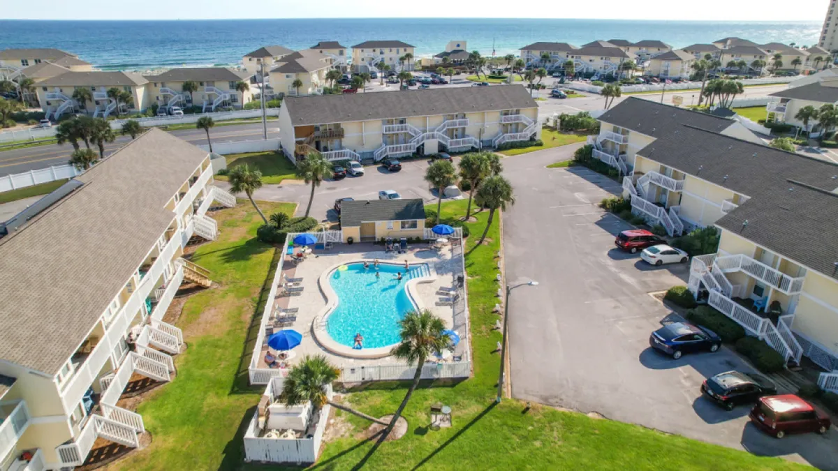 A central, sparkling swimming pool surrounded by low-rise condos with balconies, palm trees, and a glimpse of the Gulf in the background. Guests lounge poolside, while others walk towards the beach.