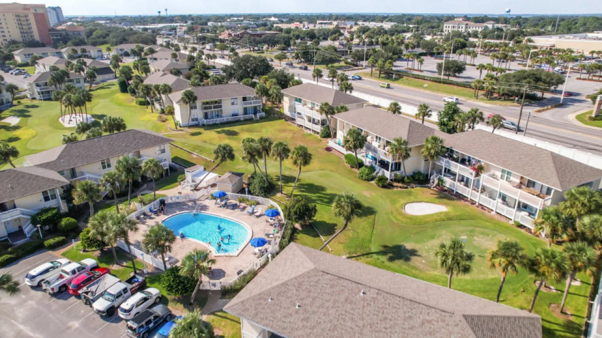 A central swimming pool with blue umbrellas is surrounded by low-rise residential buildings and balconies. Spacious green lawns with scattered palm trees create a cheerful, welcoming atmosphere. People enjoy the pool, while plenty of open lawn space suggests a pet-friendly environment and community connection.