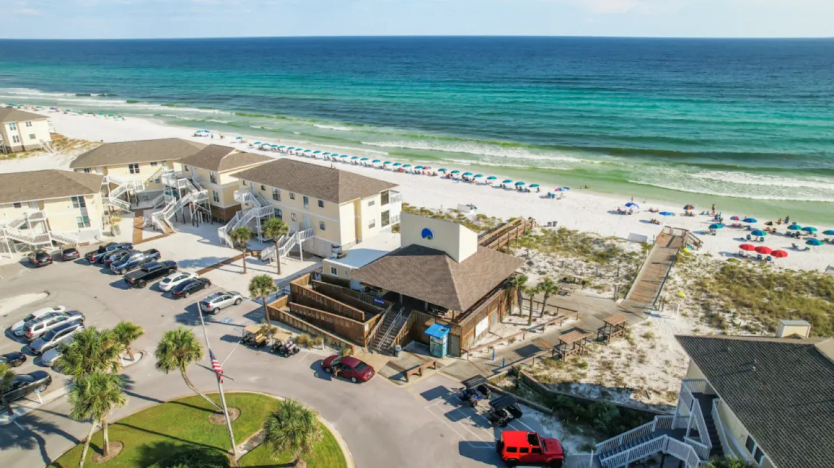 A sweeping view of Destin’s white sandy beach dotted with colorful umbrellas, with multi-story beachfront buildings and a wooden boardwalk leading directly onto the sand. The scene also shows a parking area and hints at the gentle Gulf waves.