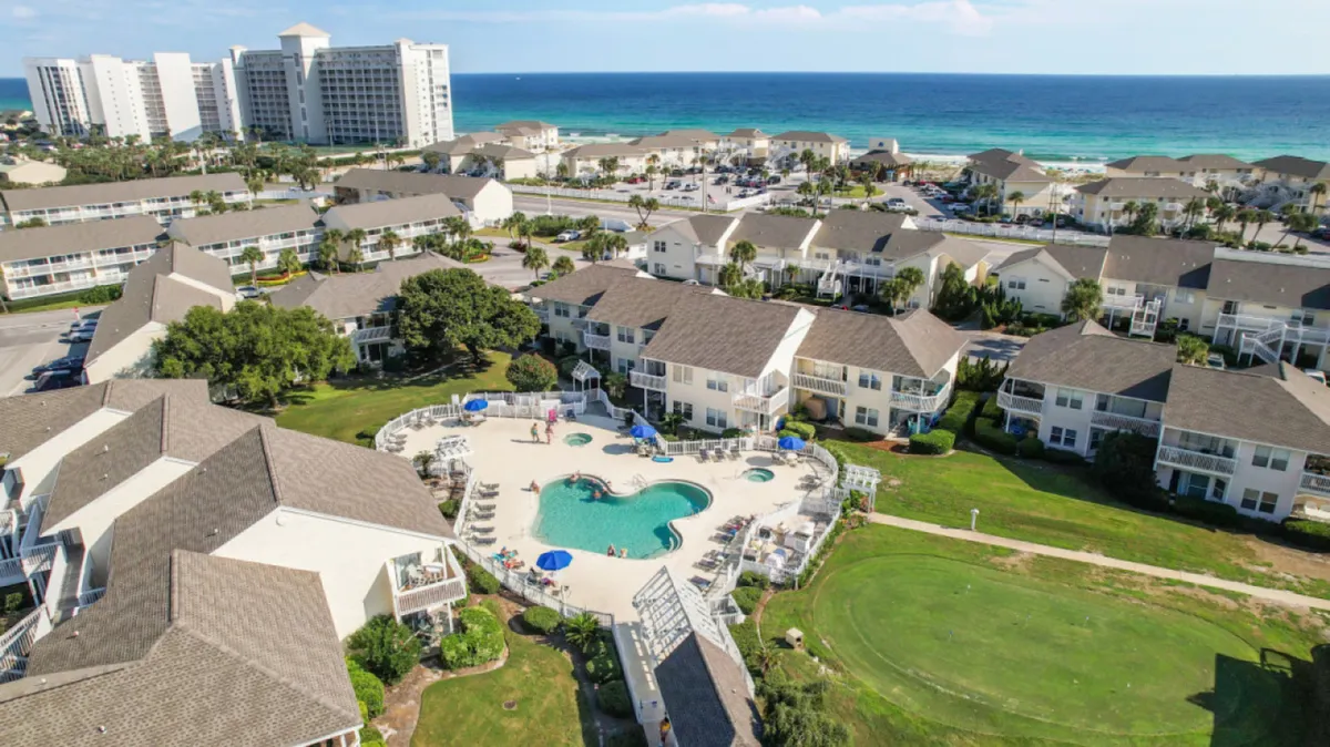 A heart-shaped swimming pool surrounded by lounge chairs and umbrellas, adjacent to a cluster of low-rise condo buildings. A large, green open lawn sits next to the pool, with the blue ocean visible in the distance. The lawn provides ample space for walks, play, and relaxation.