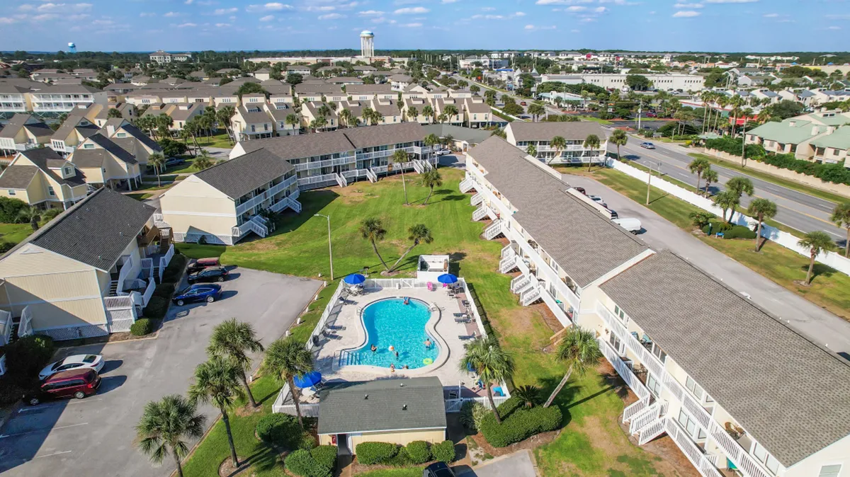 A bright, cheerful central swimming pool surrounded by lounge chairs and umbrellas, with rows of multi-unit residential buildings and lush, grassy areas lined with palm trees. The scene includes community paths and visible green spaces perfect for pet walking and outdoor enjoyment.