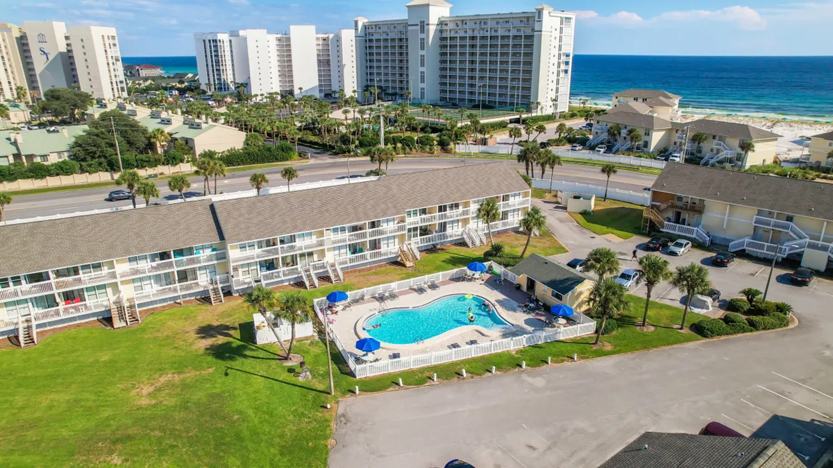 A large, sparkling swimming pool with blue umbrellas sits beside a two-story condo building and lush green lawns, with the ocean and beach visible in the background. The area is framed by palm trees, evoking a carefree, sunny vacation vibe.